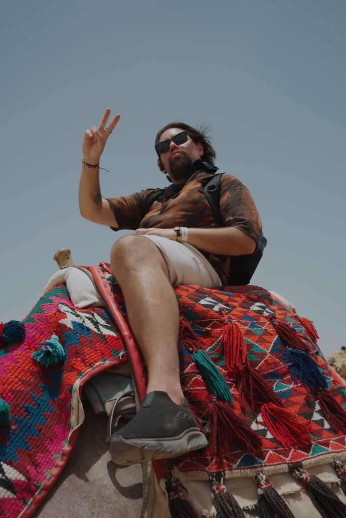 Participante sentado sobre un camello con manta tejida de colores, con el cielo del desierto al fondo.