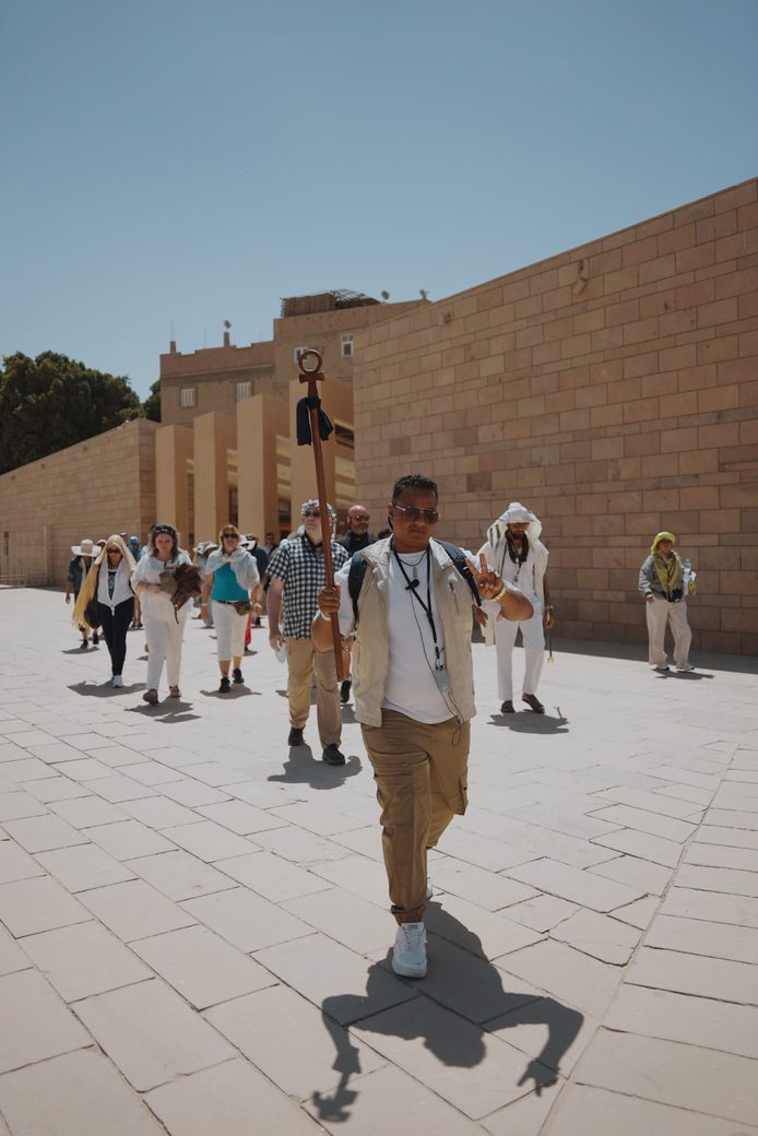 Grupo de participantes caminando junto a un muro de piedra en un recinto templario; al frente, un guía con bastón con símbolo ankh.