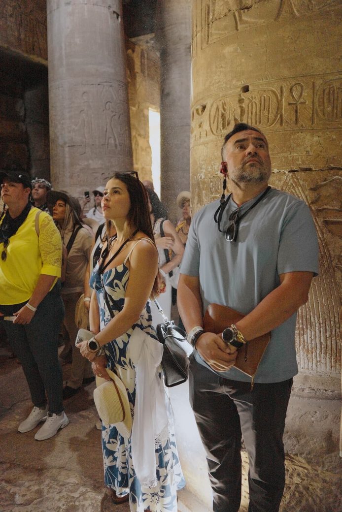 Dos participantes observan hacia arriba entre columnas con jeroglíficos iluminadas por un haz de luz.